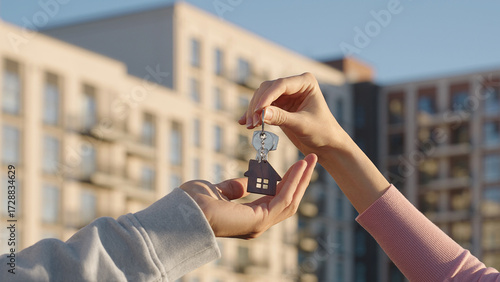Close up of female real estate owner handing over house key with home shaped keychain to young man outdoors on background of modern apartment building. New home, first time buyer, and property deal.