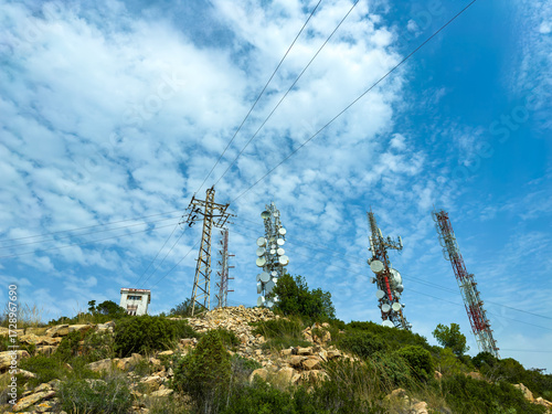 Communication towers and power lines stand tall against a blue sky with scattered clouds, captured on a sunny day in a rural area