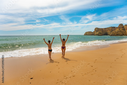 Two people run joyfully along a sandy Algarve beach, arms raised in excitement. The ocean waves crash gently as they head towards the water, embracing the freedom of the coast