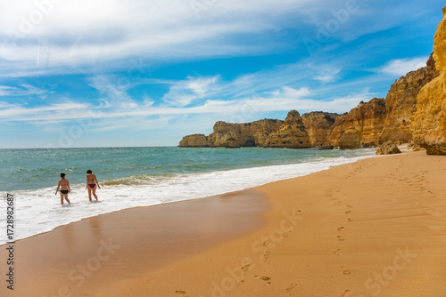 Two people run joyfully along a sandy Algarve beach, arms raised in excitement. The ocean waves crash gently as they head towards the water, embracing the freedom of the coast