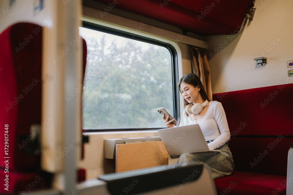 Fototapeta premium Smiling young woman traveler with suitcase and headphones around her neck holding a smartphone at the train station.