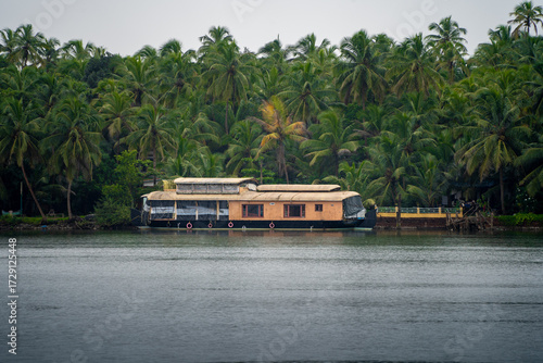 Traditional houseboat with cane bamboo roof floating away in the rain in a glassy smooth backwater in Bekal Kovalam kerela with trees in the distance showing popular tourist activity