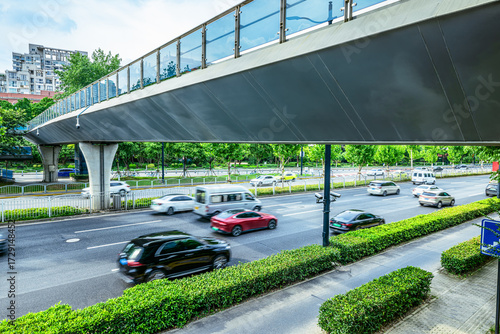 City traffic with motion blurred cars driving on a multi-lane road under a pedestrian overpass during the day.