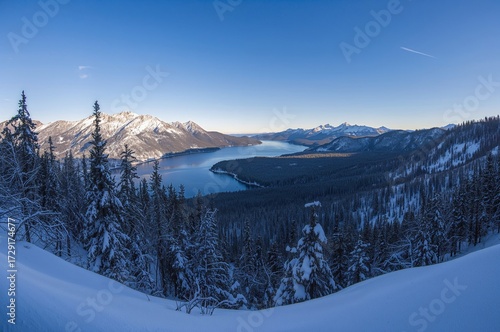 A wide-angle shot of a mountain lake encircled by snowy pine-covered slopes during winter