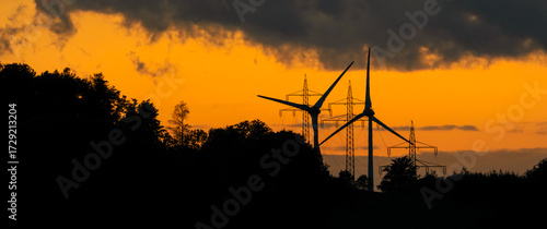 Wind turbines and power pylons against the rain clouds and orange sunset sky