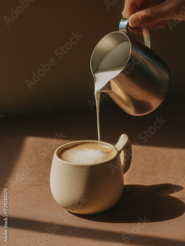 A close-up shot of a creamy coffee being prepared. A hand is pouring frothed milk from a stainless steel pitcher into a beige ceramic mug with a unique, curved handle.