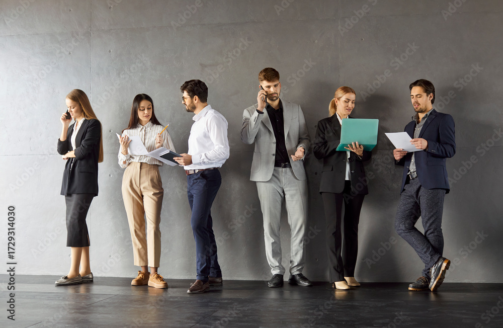 Obraz premium Portrait of confident business people in formal clothes talking on a meeting indoors. Company employees team standing in a row on dark gray wall background communicating with each other.