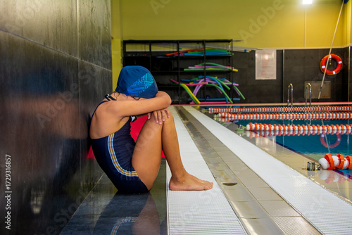 Little swimmer girl sits sadly on the floor by the pool