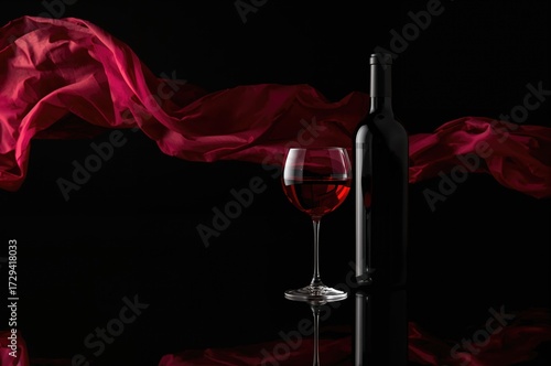 Red wine bottle and glass placed on a shiny surface with fabric waving behind. Empty space for text.