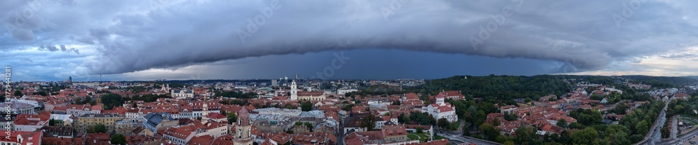Fototapeta premium Wide panoramic view of Vilnius, Lithuania, with red rooftops and historic architecture beneath a dark storm front and dramatic sky.