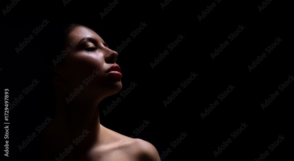 Fototapeta premium A close-up photograph of a womans face, captured in a dark environment. The woman is facing upwards, and her face is illuminated by a single light source, casting shadows across her features.