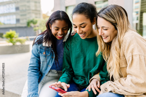 Wallpaper Mural Multiracial group of three young women is sitting close on a bench at university campus, using a mobile phone together and sharing content. Student lifestyle, social media, youth connection concept Torontodigital.ca