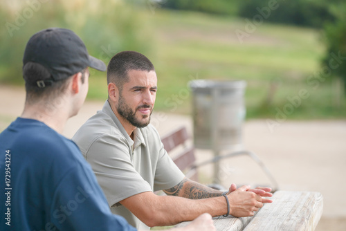 Two men are sitting on a bench in a park. One of them is wearing a hat and the other is wearing a blue shirt. They are talking to each other