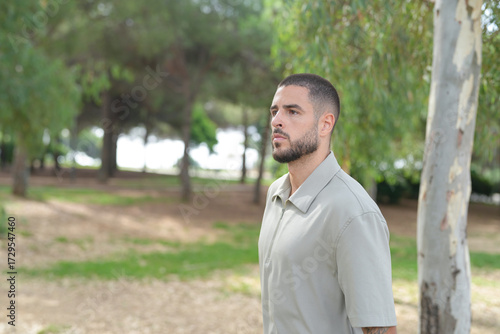 A man is standing in a park, looking at the camera. He is wearing a light-colored shirt and he is in a contemplative mood