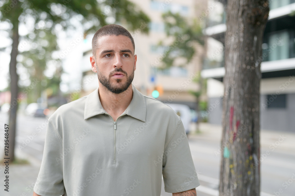 Naklejka premium A man with a shaved head and a beard stands in front of a tree. He is wearing a green shirt and he is looking at the camera