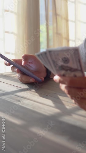 Close-up of man's hand holding smartphone and hundred dollar bills while taking notes, money financial management concept. Soft focus, Selective focus