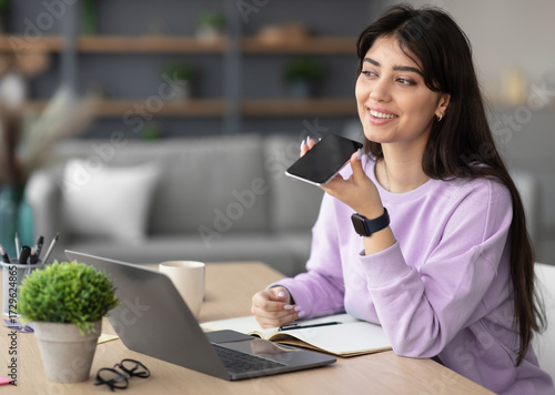 Portrait of smiling cheerful young woman sitting at desk using laptop, talking on speakerphone, dictating voice message, using online translator app or voice recognition software, virtual assistant