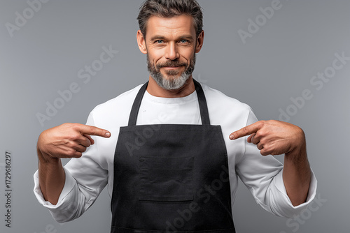 Confident, charismatic chef/barista in a clean black apron and white shirt, pointing to himself with a winning smile. Symbolizes professional readiness, service, and expertise. Mockup.
