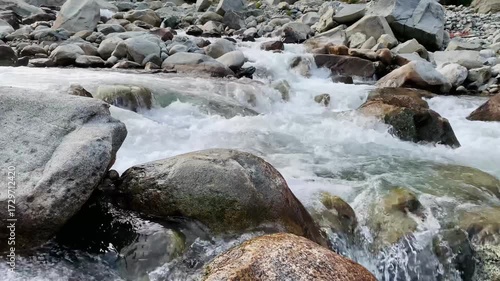 A powerful mountain stream flowing through large rocks and boulders, creating white foamy rapids. The crystal-clear water rushes with energy, showcasing the beauty of untouched nature in a rocky river