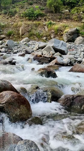 A powerful mountain stream flowing through large rocks and boulders, creating white foamy rapids. The crystal-clear water rushes with energy, showcasing the beauty of untouched nature in a rocky river