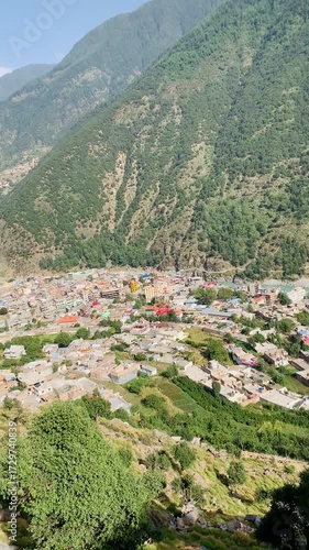 A high-angle view captures a bustling valley town nestled between towering green mountains. A vibrant turquoise river carves through the landscape.