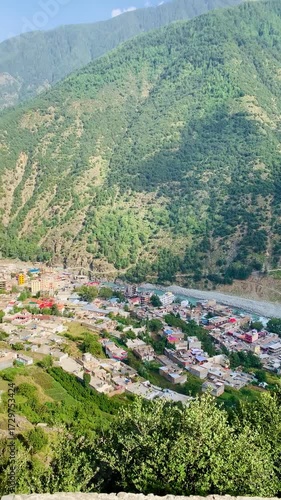 A high-angle view captures a bustling valley town nestled between towering green mountains. A vibrant turquoise river carves through the landscape.