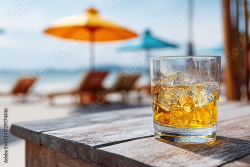 A glass of whiskey with ice on a weathered wooden table with a blurred beach scene of umbrellas and loungers in the background