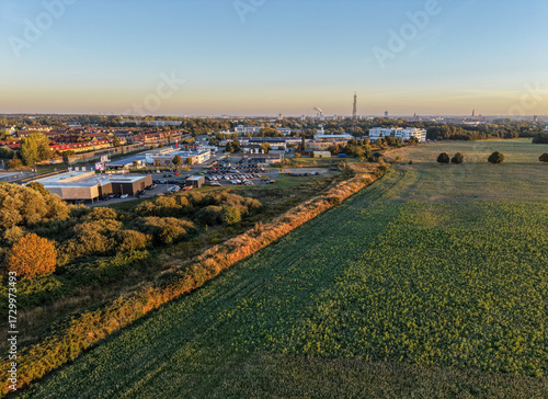 Aerial view of a town’s outskirts where industry meets open green fields, bathed in warm morning light with a distant skyline under a clear sky