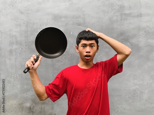 A southeast asian teenager with shocked and startled expression in a red t-shirt carries a frying pan