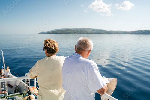 Elderly man standing on the deck of a ferry boat in Greece, looking at the calm blue Aegean Sea and the green coastline in the distance.