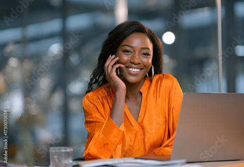 Smiling woman in orange blouse on phone with laptop