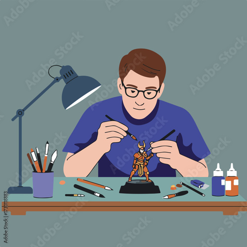 Man paints miniature figure with brushes under a lamp on a cluttered desk