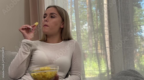 The teenager is eating chips. A teenage girl comfortably nestles on the sofa, happily eating chips from a bowl in a bright, cozy living room.