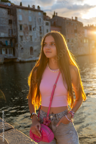 Teen girl in pink posing by the water at sunset. A stylish teenage girl with long hair stands confidently by the sea during golden hour, backlit by the warm setting sun.