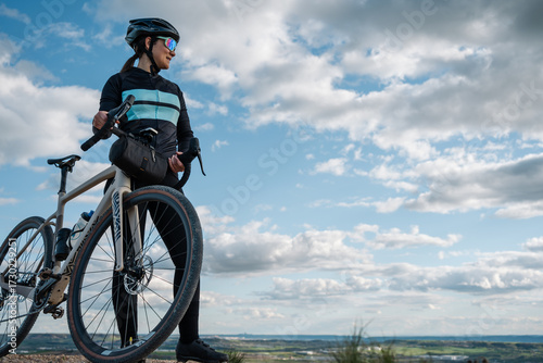 Young female cyclist resting with gravel bike on hilltop enjoying scenic view