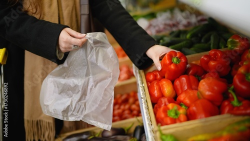 Woman's hands picking fresh red bell pepper from a produce display into a plastic bag at a supermarket