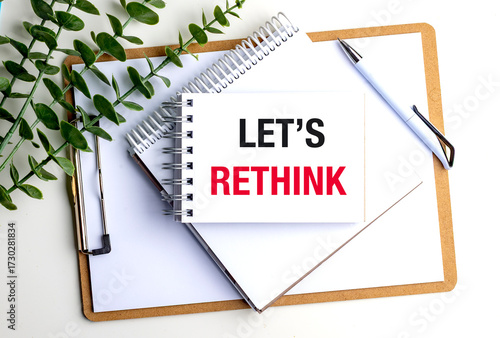 Notebook with the words let is rethink on clipboard with white pen, spiral notepad and green leaves