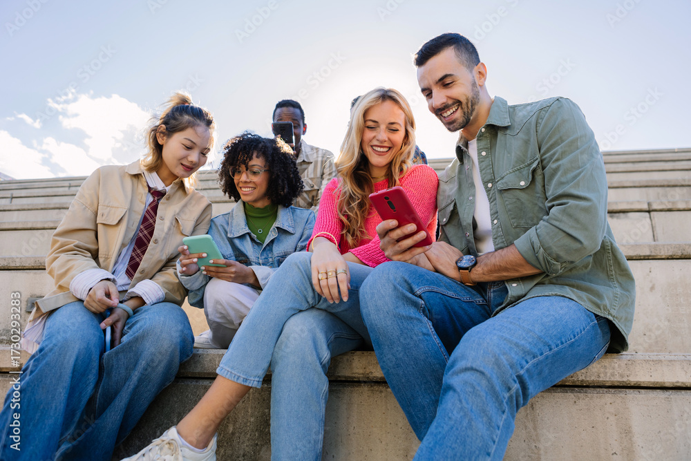 Fototapeta premium Group of young multiracial students sharing a moment outdoors while looking at a smart phone screen. Youth community, friendship and lifestyle concept.