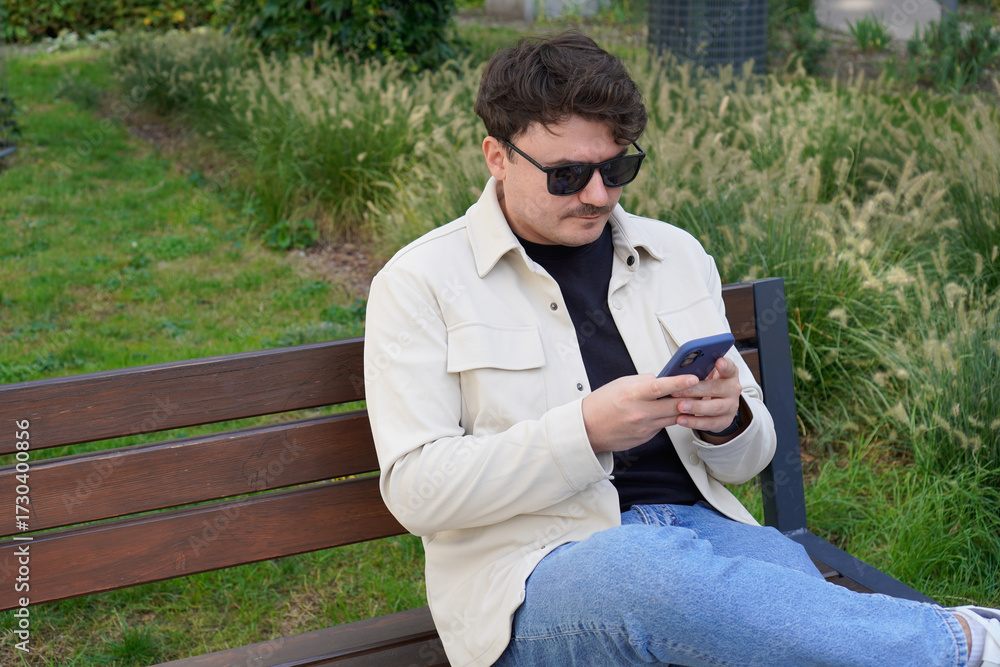 Fototapeta premium A Caucasian man with glasses sits on a bench and types a message on his phone