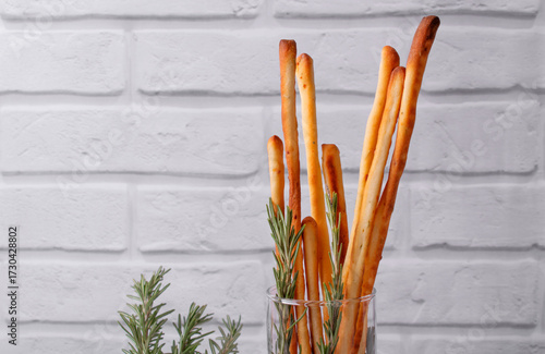 Grissini made of wheat flour with parmesan, rosemary and olive oil served in a glass. Breadsticks, Italian crispy snack, against the grey brick wall. Copy space
