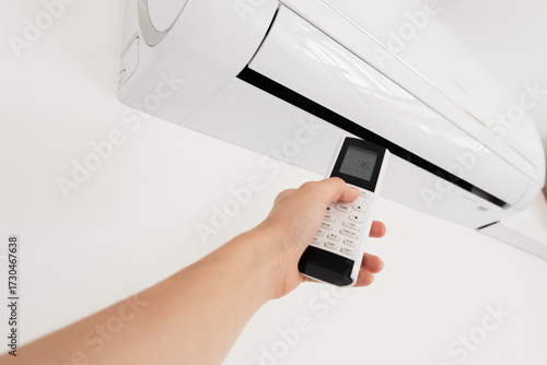 A hand holding a remote control aimed at a wall-mounted air conditioner. The air conditioner is white and modern in design.