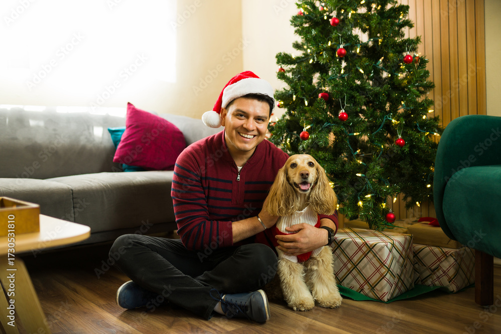 Obraz premium Portrait of a Hispanic man hugging his dog wearing christmas clothes by the tree