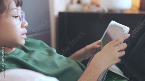 Selective focus of Asian kid girl, playing game on tablet with concentration. Concepts of children technology use, online learning, screen time, digital lifestyle, and modern childhood education.