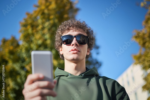 Young visually impaired man in dark glasses and white sweater using a smartphone application for blind people in the city. Mobile phone technology for vision loss persons. Low vision individuals.