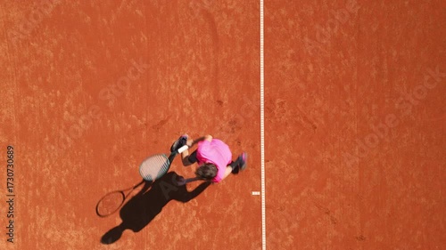 Tennis player in pink shirt practices on clay court, dynamic movement and focus captured from aerial shot, highlighting shadow and dedication to skill development.