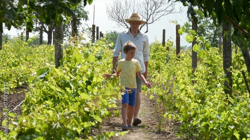 Young Man in Straw Hat Walks with Son after Work with Pitchfork in Garden and Vineyard at Summer. Agriculture Concept and Agrotourism with Rural Lifestyle. Farmer