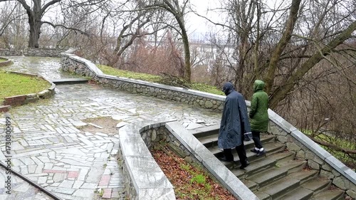 Two people in raincoats ascend wet stone stairs in a park on an overcast, chilly day. Bare trees and stone walls frame the path, hinting at a quiet, contemplative journey through nature.