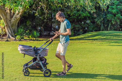 Father pushing a baby stroller on a grassy field while looking at his phone. Concept of modern parenting, multitasking, technology in daily life, and work-life balance