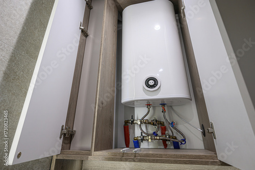 A white hot water heater sits inside a cabinet with open doors, revealing pipes