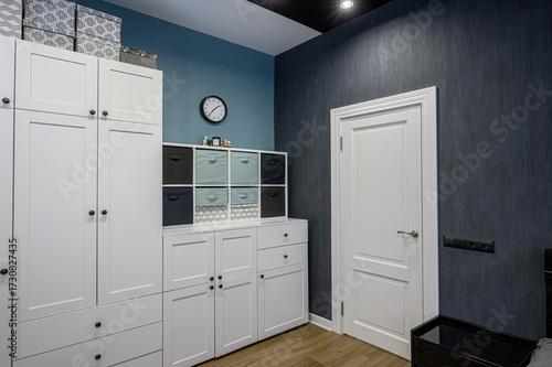 Bild auf Leinwand cozy modern wardrobe nook with white IKEA-style cabinets, gray-blue textured walls, colorful storage bins, wall clock, potted plant, white door, and wooden floor
